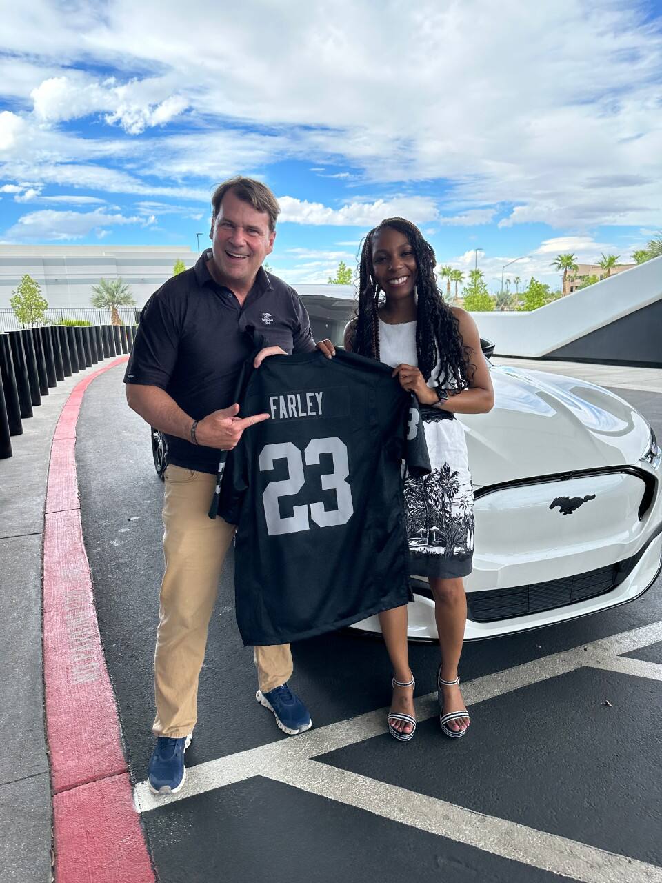 A man and woman stand in front of a white Ford mustang. They hold a jersey that says "Farley 23" on the back.